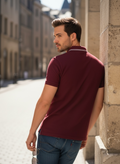Man wearing a burgundy polo shirt and blue jeans leaning against a stone wall.