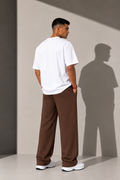 A man standing with his back to the camera wearing a white t-shirt and brown wide-leg pants, set against a minimalist gray wall with a prominent cast shadow.