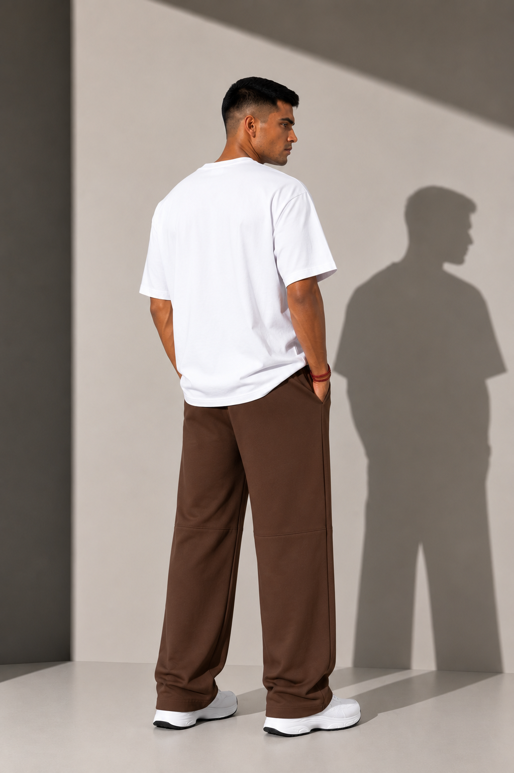 A man standing with his back to the camera wearing a white t-shirt and brown wide-leg pants, set against a minimalist gray wall with a prominent cast shadow.