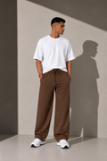 A man wearing a white crewneck t-shirt and brown drawstring wide-leg sweatpants, standing in a minimalist studio with dramatic shadows.