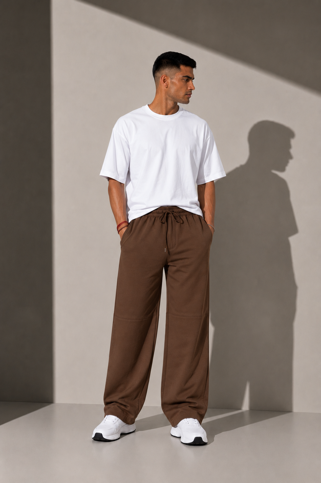 A man wearing a white crewneck t-shirt and brown drawstring wide-leg sweatpants, standing in a minimalist studio with dramatic shadows.