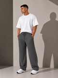 A man wearing a white crewneck t-shirt and dark gray drawstring sweatpants, standing in a studio with a strong cast shadow on the wall behind him.