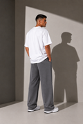 A man stands with his back to the camera wearing a white t-shirt and loose gray sweatpants, standing in a minimalist studio with dramatic side lighting that casts a long shadow on the wall.