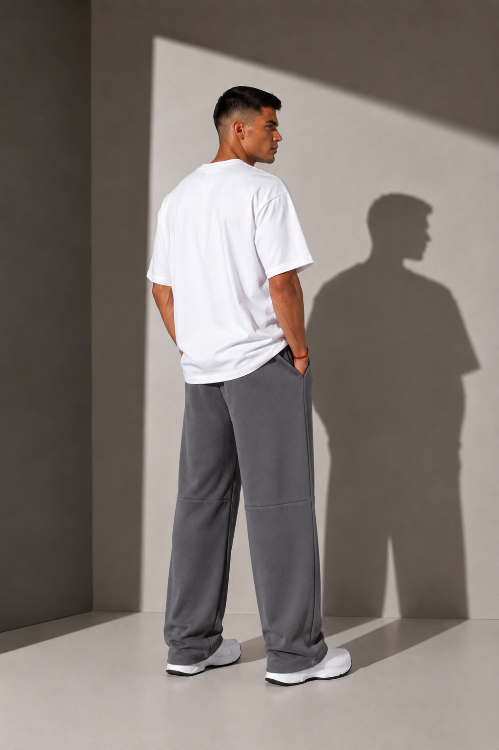 A man stands with his back to the camera wearing a white t-shirt and loose gray sweatpants, standing in a minimalist studio with dramatic side lighting that casts a long shadow on the wall.
