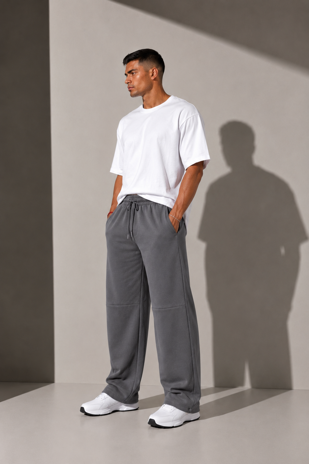 A man wearing a white crewneck t-shirt and gray drawstring sweatpants, standing in a studio with dramatic side lighting and a sharp shadow on a light gray background.